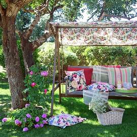 Different Patterned Scatter Cushions On Rustic Garden Swing Seat Under Shady Tree With Picnic Basket On Ground by Winfried Heinze