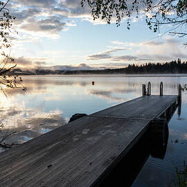Dock By Calm Lake And Tree During Sunrise With Reflection Of Clouds by Cavan Images