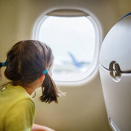 Little Girl With Pigtails Sits On Plane Staring Out Window On Tarmac by Cavan Images