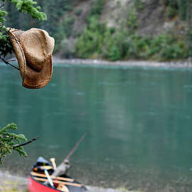 Wet Straw Hat Hangs From A Tree, In The Background A Canoe On The Shore, Break At The Yukon River, Yukon, Canada by Myriam Brunner