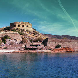 Crete, Greece Wide angle view of Spinalonga unhabited island with a 16th century venetian fortress and the ruins of a formar leper colony against cloudy blue sky and blue water in the foreground