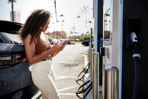 Wall Art - Photograph - Young Woman Using Phone Leaning On Electric Car At Charging Station by Cavan Images / Jack Jeffries
