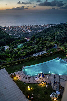 Wall Art - Photograph - Evening Celebration by the Pool - A vibrant wedding reception scene by a pool illuminated with colorful lights against mount vesuvius volcano surrounded with dramatic clouds by Arpan Bhatia