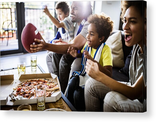 Unhealthy Eating Acrylic Print featuring the photograph Cheerful black family cheering while watching rugby match on TV at home. by Skynesher