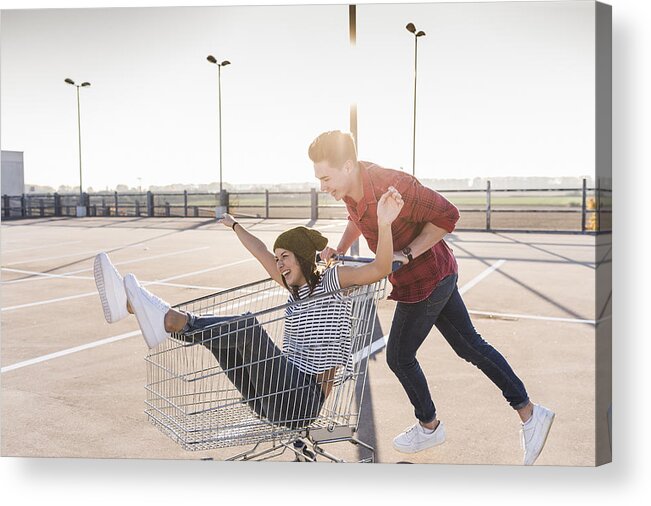 Young Men Acrylic Print featuring the photograph Playful young couple with shopping cart on parking level #1 by Westend61