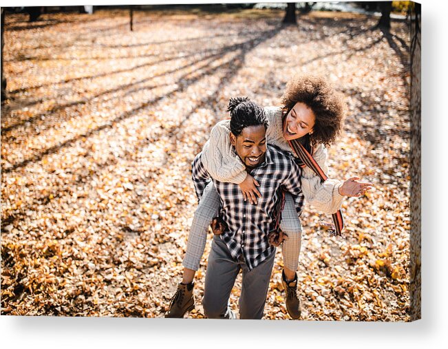 Young Men Acrylic Print featuring the photograph Cheerful African American couple piggybacking in autumn at the park. by Skynesher