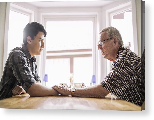 Young Men Acrylic Print featuring the photograph Side view of grandfather consoling grandson at table by Maskot