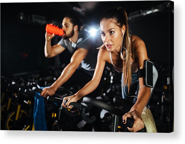 Young Men Acrylic Print featuring the photograph Two young people exercising on exercise bike at gym by EmirMemedovski