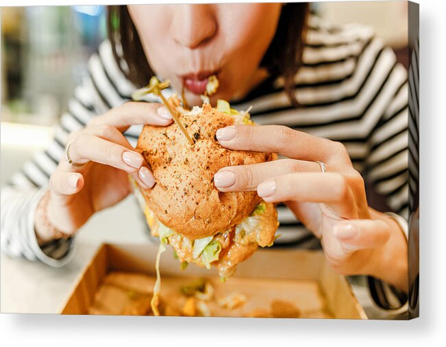 Unhealthy Eating Acrylic Print featuring the photograph Woman eating a hamburger in modern fastfood cafe, lunch concept by Frantic00