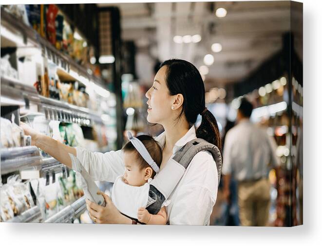 Toddler Canvas Print featuring the photograph Beautiful Asian woman carrying cute baby girl shopping for organic green marketing products in grocery store by D3sign