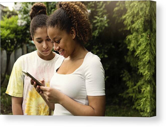 Sibling Canvas Print featuring the photograph Mixed-race teenage sisters looking at mobile phone in backyard. #2 by Martinedoucet