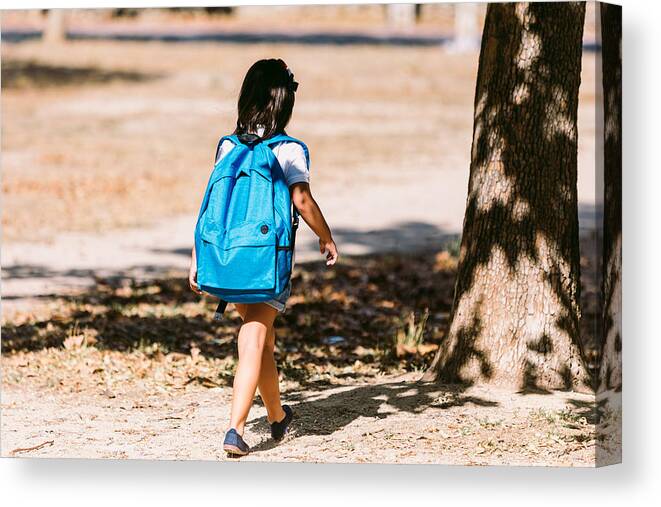 4-5 Years Canvas Print featuring the photograph A black-haired girl with a blue backpack, turned her back, walks through a park in the direction of school. Concept of education and back to school. by DBenitostock