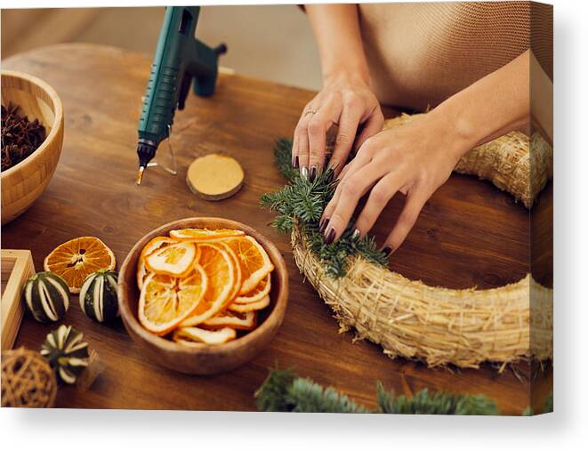 People Canvas Print featuring the photograph Close-up of unrecognizable woman with manicure standing at wooden table in workshop and using hot glue while attaching fir tree twigs to wreath base by Mediaphotos