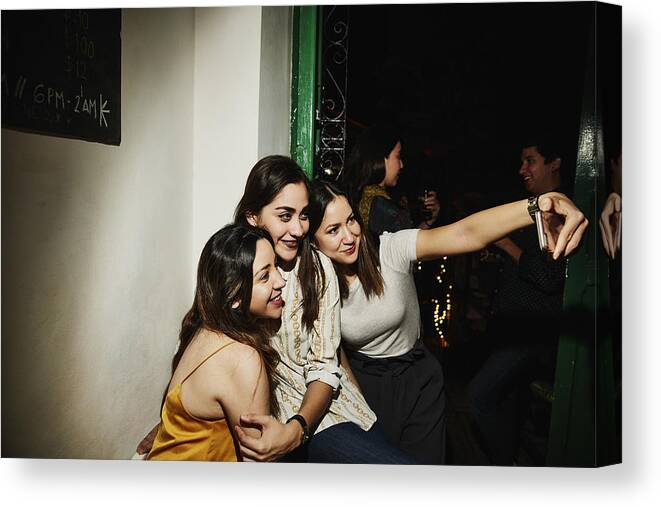 Adults Only Canvas Print featuring the photograph Smiling female friends taking selfie with smart phone while hanging out in night club by Thomas Barwick