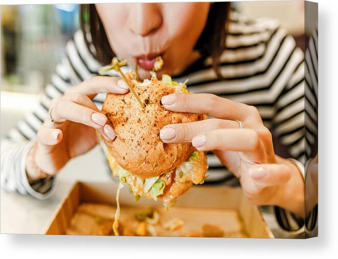 Unhealthy Eating Canvas Print featuring the photograph Woman eating a hamburger in modern fastfood cafe, lunch concept by Frantic00