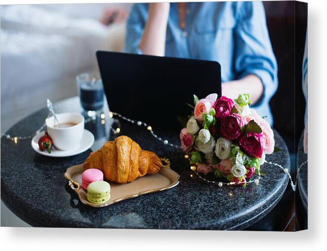 New Business Canvas Print featuring the photograph Young woman freelancer in blue shirt working on laptop during lunch with coffee, croissant, macaroons and flowers on table by Alexa-photo