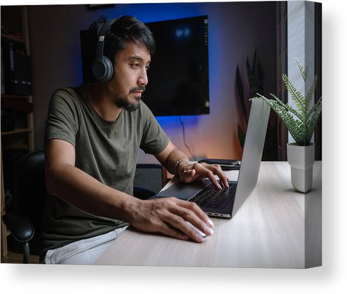 Young Men Canvas Print featuring the photograph Millennial man playing computer game on laptop at home. by Staticnak1983
