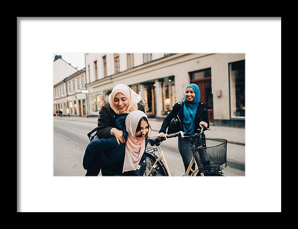 Three Quarter Length Framed Print featuring the photograph Cheerful teenage girl giving young woman piggyback by friend walking with bicycle on street in city by Maskot