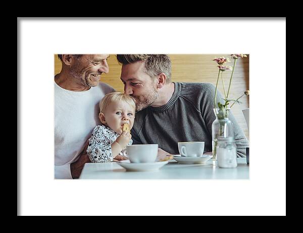 Mature Adult Framed Print featuring the photograph Gay couple cuddling with their baby in cafe by Westend61
