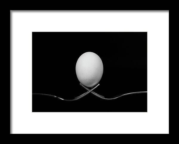 Egg Framed Print featuring the photograph White egg resting on two metal and shiny forks on a black backg by Michalakis Ppalis