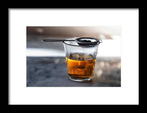 Black Tea Framed Print featuring the photograph Glass filled with hot tea from a strainer filled with tea leaves on top on a gray marble tabletop. by Emreturanphoto