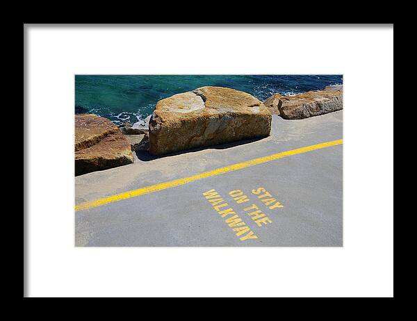 Water's Edge Framed Print featuring the photograph 'Stay on the Walkway' sign stencilled in yellow paint on the concrete walkway of the Southern Break Wall at Coffs Harbour, New South Wales, Australia by Simon McGill