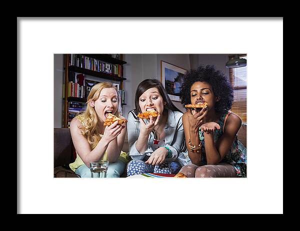 People Framed Print featuring the photograph Women biting into pizza sitting together on sofa. by Betsie Van Der Meer