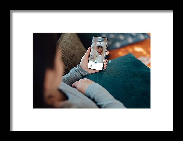 People Framed Print featuring the photograph Young mother using baby monitor app on her smart phone while sitting in the living room by Oscar Wong