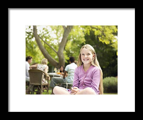 Cardigan Sweater Framed Print featuring the photograph Young Smiling Girl Sits Cross-Legged in a Garden, People at a Table in the Background by Digital Vision.
