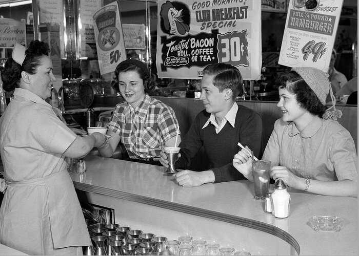 B&w Black And White 1950s 1960s Adult Americana Beverage Boy Breakfast Children Counter Countertop Date Dessert Diner Drink Eat Female Food Fountain Friendship Girl Hamburger Happy Hat Ice Cream Indoors Junk Food Juveniles Kids Lifestyle Lunch Male Malt Menu Mid-adult Woman Milkshake People Service Sign Smile Snack Food Soda Soda Fountain Soda Jerk Teenage Boy Teenage Girl Teenager Teen Togetherness Waitress Woman Young Youngster Youth Retro Vintage Nostalgia Nostalgic Old Fashioned Old Fashion Greeting Card featuring the photograph 1950s Smiling Woman Waitress Serving Two Teenage Girls And One Boy At Diner Counter by Panoramic Images