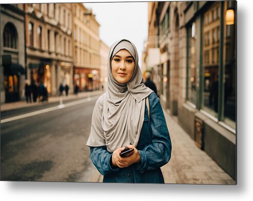 Asian And Indian Ethnicities Metal Print featuring the photograph Portrait of confident young woman wearing hijab standing with mobile phone on sidewalk in city by Maskot