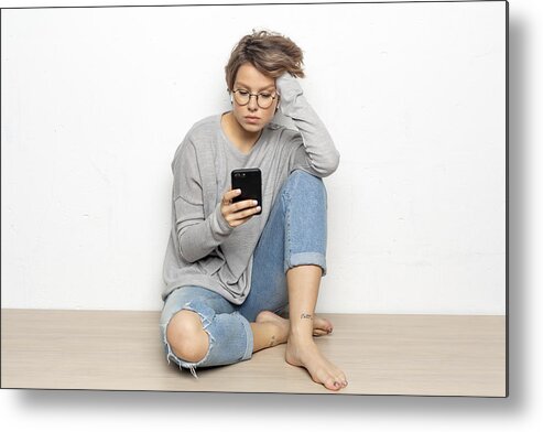 Sitting On Floor Metal Print featuring the photograph Portrait of young woman with wireless earphones sitting on the floor looking at cell phone by Westend61