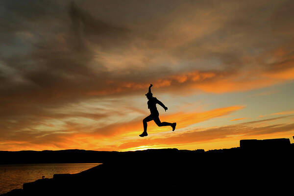 Environment Poster featuring the photograph Silhouette Of Man Jumping With Cap In The Sunset #1 by Cavan Images