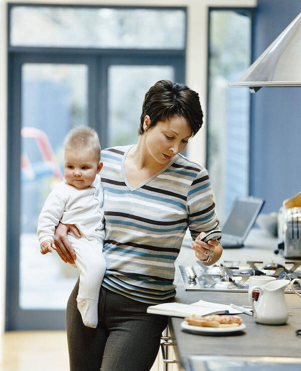 Breakfast Art Print featuring the photograph Woman Stands by a Kitchen Counter Holding Her Baby and Dialing Her Mobile Phone by Digital Vision.