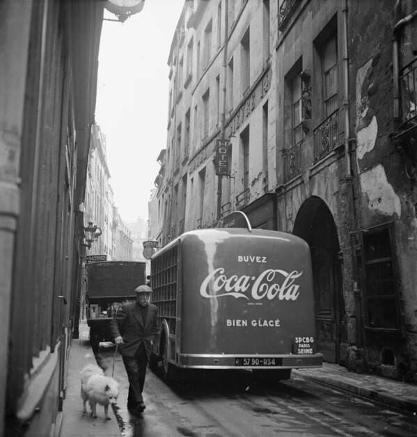 Advertisement Art Print featuring the photograph A man walks his dog beside a bus with a large advertisement for Coca Cola on its back. The ad reads Drink Coca Cola well chilled. by Mark Kauffman