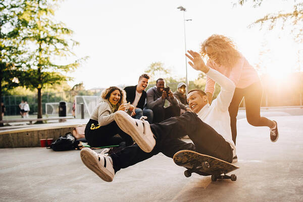 Young Men Art Print featuring the photograph Laughing friends photographing man falling from skateboard while woman pushing him at park by Maskot