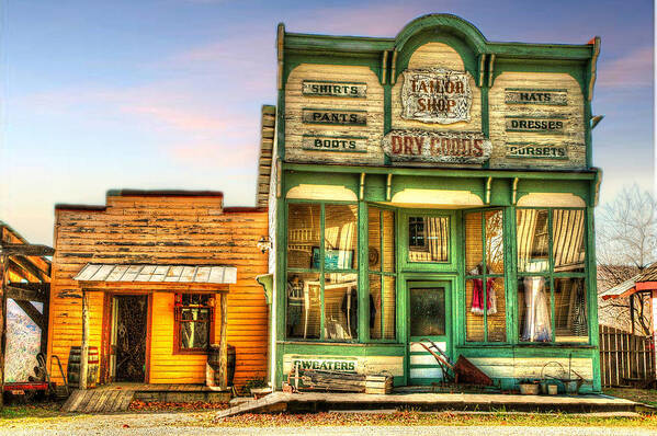 Virginia City Dry Good Store. Merchantile. Vintage Archetecture. Buildings. Sled. Wheelbarrow. Vintage Dresses. Wool Spinners. Lady Clothing. Children Clothing. Mens Clothing. Photography. Fine Art. Canvas. Texture. Hdr. Print. Digital Art. Poster. Greeting Card. City Scene. Cloudy Skies. Fence. Art Print featuring the photograph Virginia City Dry Goods by Mary Timman