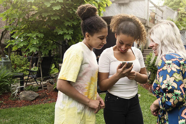 Sibling Art Print featuring the photograph Mixed-race sisters looking at mobile phone with mother in backyard. by Martinedoucet
