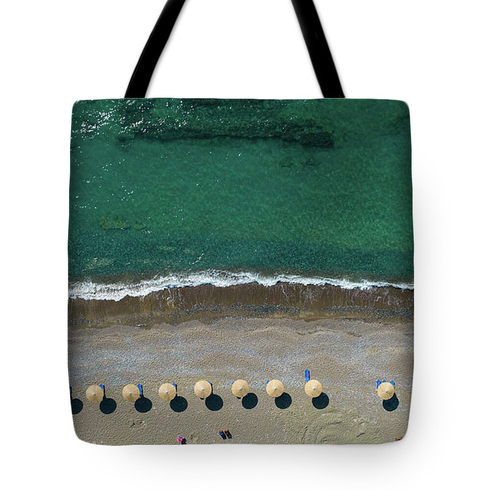 Summertime Tote Bag featuring the photograph Aerial view from a flying drone of beach umbrellas in a row on an empty beach with braking waves. by Michalakis Ppalis
