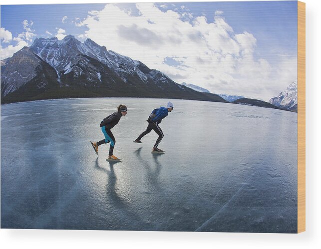 Following Wood Print featuring the photograph A man leads a woman on a winter speed skating adventure on Lake Minnewanka in Banff National Park, Alberta, Canada. #1 by GibsonPictures