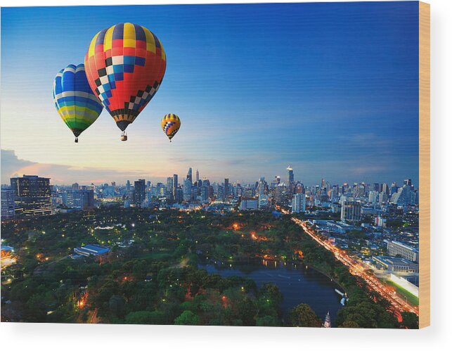 Skyline Trail Wood Print featuring the photograph Hot air balloons fly over cityscape at sunset background by Busakorn Pongparnit