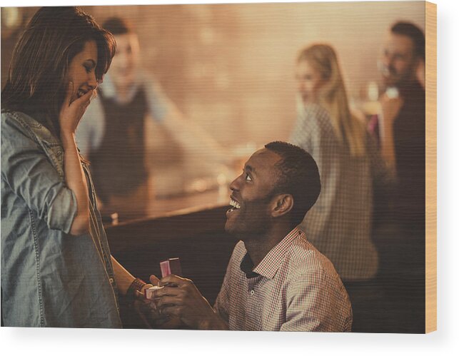 Heterosexual Couple Wood Print featuring the photograph Happy African American man proposing to his girlfriend in a bar. by Skynesher