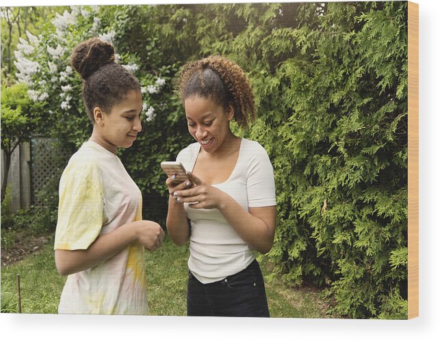 Sibling Wood Print featuring the photograph Mixed-race teenage sisters looking at mobile phone in backyard. by Martinedoucet