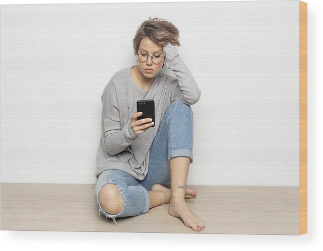 Sitting On Floor Wood Print featuring the photograph Portrait of young woman with wireless earphones sitting on the floor looking at cell phone by Westend61