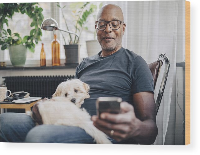 Pets Wood Print featuring the photograph Smiling retired senior male using smart phone while sitting with dog in room at home by Maskot