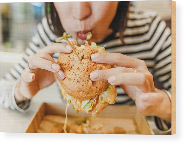 Unhealthy Eating Wood Print featuring the photograph Woman eating a hamburger in modern fastfood cafe, lunch concept by Frantic00
