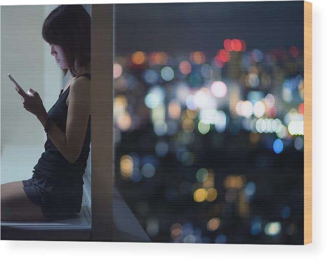 Apartment Wood Print featuring the photograph Japanese woman sitting with back to corner high-rise window using smartphone by Benjamin Torode