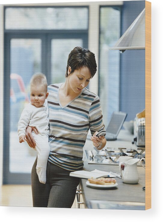 Breakfast Wood Print featuring the photograph Woman Stands by a Kitchen Counter Holding Her Baby and Dialing Her Mobile Phone by Digital Vision.