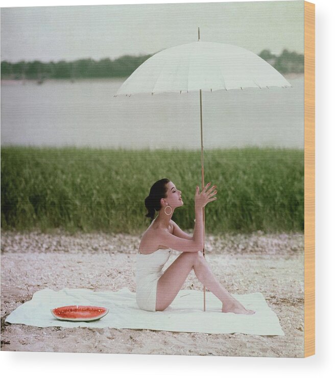 One Person People Outdoors Daytime Fashion Model Model Fashion Clothing 25-29 Years Young Adult 20s Adult Female Young Woman Young Adult Woman Umbrella Holding Brunette Dark Hair Ponytail Earrings Jewelry Watermelon Towel Beach Coastline Sand Sea Water Summer Seasons Sitting Eyes Closed Barbara Mullen Swimsuit Sunbathing Leisure Relaxation Grass Barefoot White #condenastglamourphotograph July 1st 1954 Wood Print featuring the photograph Model Barbara Mullen In A White Swimsuit by Frances McLaughlin-Gill