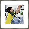 Boy (6-8) Putting Bowls Away In Kitchen Cupboard, Side View Framed Print